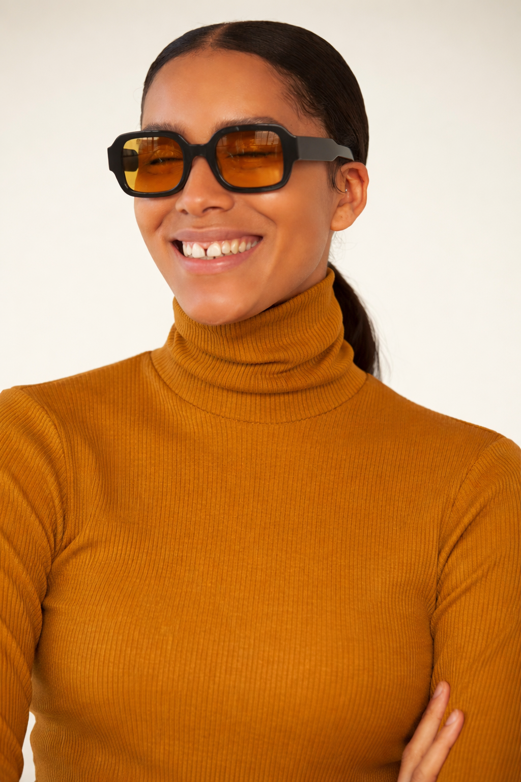 Woman wearing sunglasses and a mustard yellow turtleneck sweater against a plain background