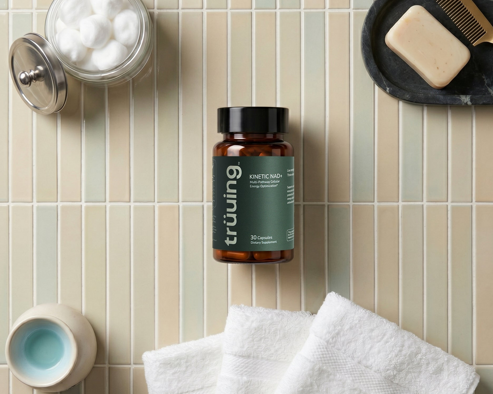 Kinetic_NAD bottle of trüuing Shitake Mucor supplement capsules is displayed in a styled bathroom flat-lay setting on a beige and green striped tile background, surrounded by towels, a jar of cotton balls, a soap dish with a comb, and a small cup.